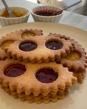A stack of lunette cookies with jam windows on a plate, with bowls of strawberry and apricot jam behind