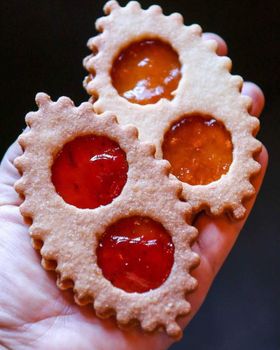 A hand holds four lunette cookies with strawberry and apricot jam windows against a dark background