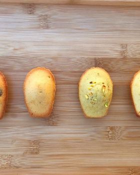 Four madeleines side by side on a wooden board: chocolate chip, vanilla, pistachio and lemon