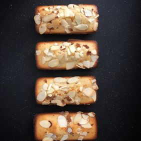 Four rectangular almond financiers topped with sliced almonds on a black surface