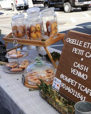 Glass jars of madeleines, pastries under a glass dome and a letterboard reading Giselle et Mathieu Petit Cafe on a market table