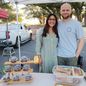 A woman and a man stand arm in arm behind the market table with boxed pastries and a letterboard sign