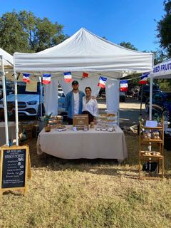 Two people stand under the GM Petit Cafe tent behind a table of pastries at the Pedernales Farmers Market