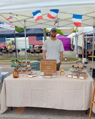A man in a cap stands behind the GM Petit Cafe market table under a tent with French flag bunting