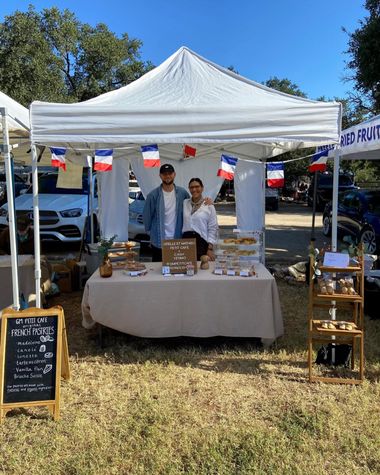 Two people under a white market tent with French flag bunting beside a table and shelves of pastries