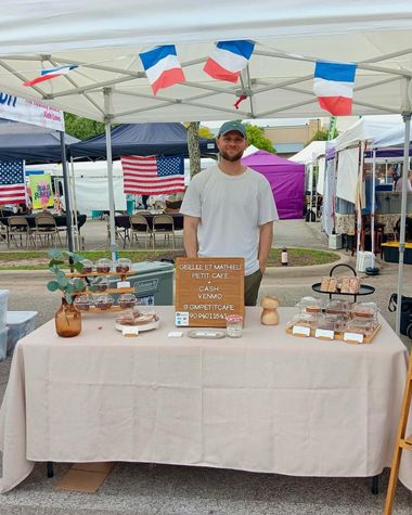 A man in a cap stands behind the GM Petit Cafe table under a canopy with French flag bunting at Wolf Ranch Farmers Market