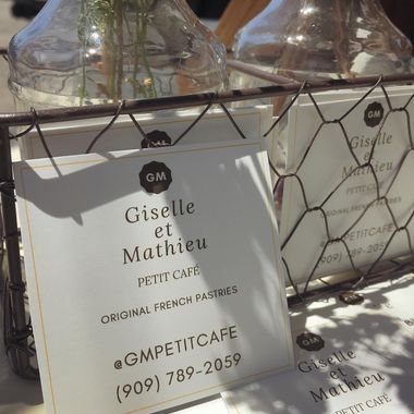 The first GM Petit Cafe business cards, reading Giselle et Mathieu Petit Cafe, propped in a wire rack beside glass bottles