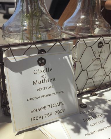 The first GM Petit Cafe business cards, reading Giselle et Mathieu Petit Cafe, propped in a wire rack beside glass bottles