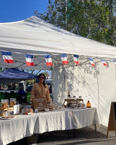 A woman stands behind the GM Petit Cafe table under a white canopy with French flag bunting