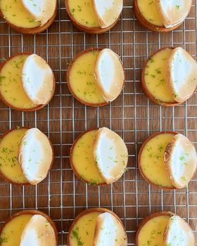 A dozen lemon meringue tarts with toasted meringue and lime zest on a wire rack, seen from above